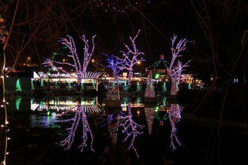 Colorful holiday lights illuminate trees and a gazebo, reflecting in a calm water surface at night.