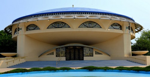 A modern, circular building with a blue dome and decorative windows, surrounded by greenery and a small water feature.