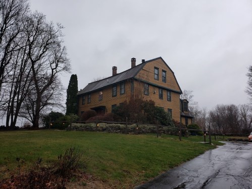 A large, two-story yellow house with a sloped roof, surrounded by trees and a wet driveway on a cloudy day.