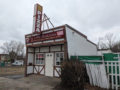 A small pizza restaurant with a brick oven sign, featuring a white exterior and a green tarp covering part of the building.