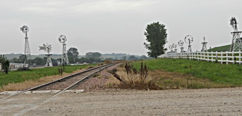 A rural scene featuring a winding railway track, green hills, and several windmills under a cloudy sky.