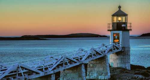 A white lighthouse stands on a rocky pier at sunset, with calm waters and distant hills in the background.