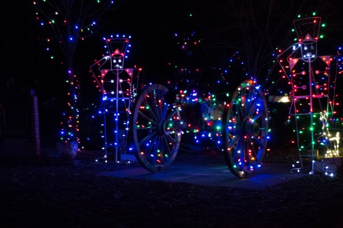 Colorful holiday lights illuminate two toy soldier figures beside a decorated wagon in a nighttime scene.