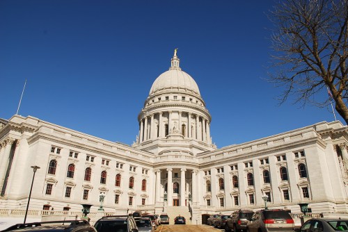 The Wisconsin State Capitol building with a large dome, surrounded by clear blue sky and parked cars in front.