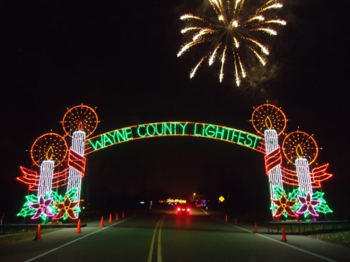 Colorful holiday lights form an archway reading "Wayne County Lightfest," with fireworks in the night sky above.