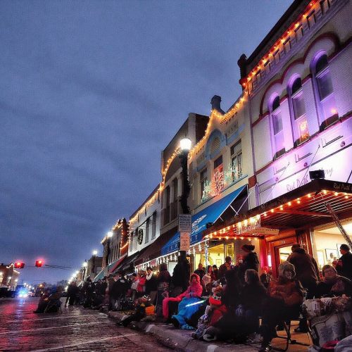 A festive street scene at dusk, with people gathered along a decorated storefront, illuminated by holiday lights.