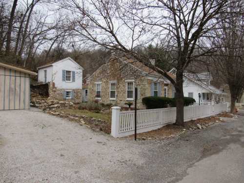 A stone house with a white fence, surrounded by trees, on a gravel driveway in a rural setting.