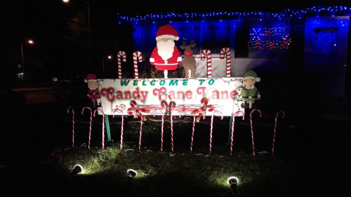 A festive sign reading "Welcome to Candy Cane Lane," decorated with candy canes, Santa, and holiday lights.