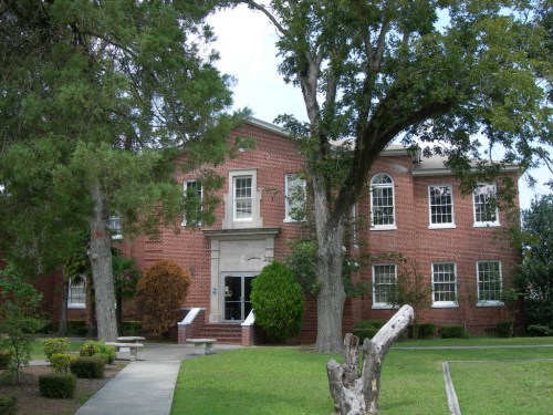 A brick building surrounded by trees and greenery, featuring large windows and a welcoming entrance.