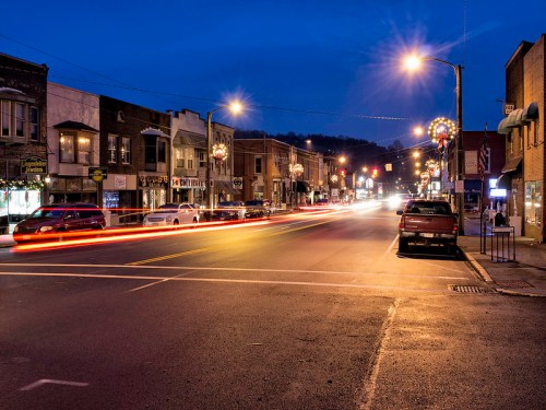 A quiet street at dusk, lined with shops and festive lights, with car headlights creating streaks of light.