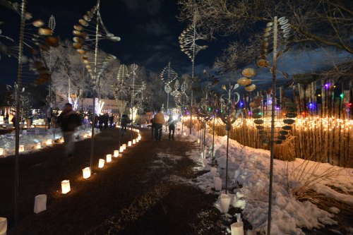 A snowy path lined with glowing lanterns and artistic sculptures, illuminated by colorful lights at night.