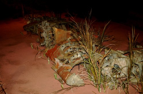 A rusted aircraft wreckage partially covered by grass on a sandy surface.