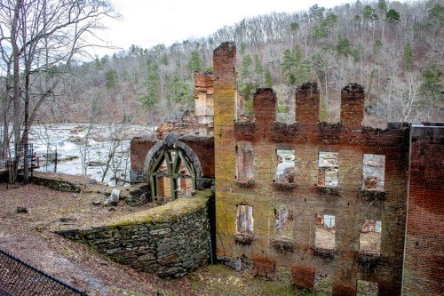 Ruins of a brick building with moss-covered walls, surrounded by trees and a river in the background.