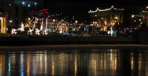 A festive display of colorful lights reflecting on water at night, with decorated trees and buildings in the background.