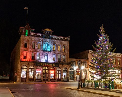 Historic building with colorful lights at night, next to a decorated Christmas tree in a quiet town square.