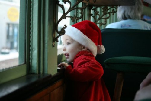 A child in a Santa hat gazes out a window, looking curious and excited.