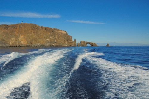 A scenic view of a rocky coastline with a natural arch, waves trailing behind a boat in calm blue waters.