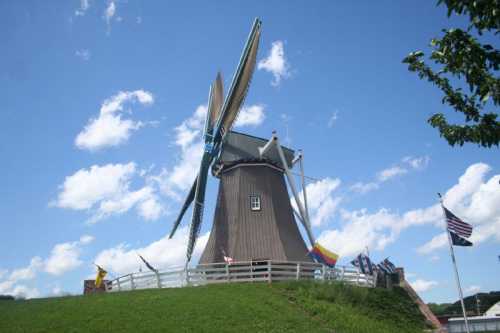 A tall windmill with large blades, surrounded by green grass and flags, under a blue sky with fluffy clouds.