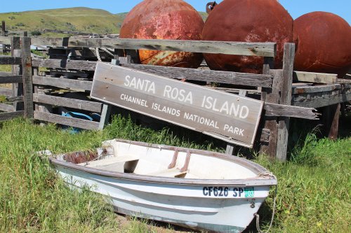 A small boat rests in front of a weathered sign for Santa Rosa Island, Channel Islands National Park, with large buoys nearby.