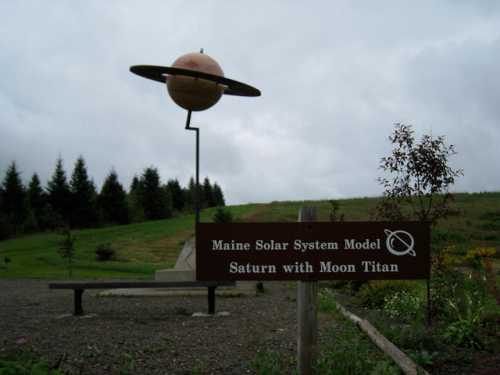 A large model of Saturn with its moon Titan, set in a grassy area with trees and a sign describing the installation.