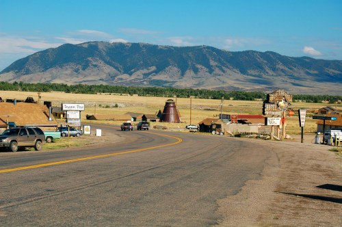 A scenic view of a rural road leading to a small town, with mountains in the background and a clear blue sky.