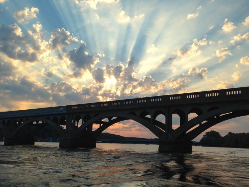 Silhouette of a bridge against a vibrant sunset with rays of light breaking through clouds over water.