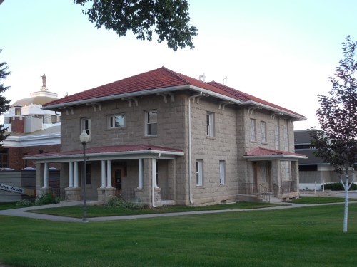 Historic stone building with a red tile roof, surrounded by green grass and trees, in a serene outdoor setting.