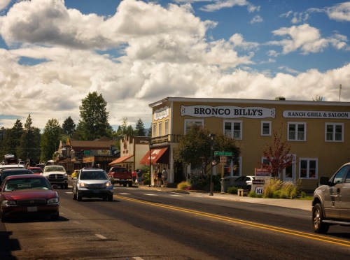 A street view featuring Bronco Billy's Ranch Grill & Saloon, with cars parked and shops lining the road under a cloudy sky.