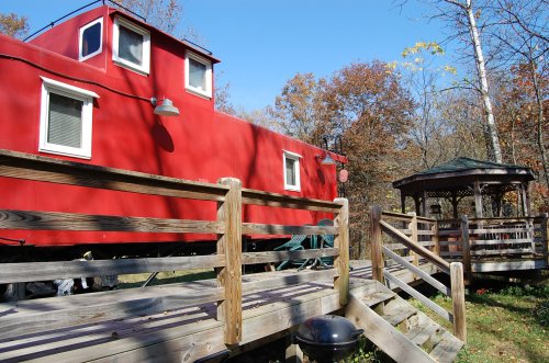 A red caboose sits beside a wooden gazebo, surrounded by trees with autumn foliage under a clear blue sky.