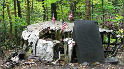 A weathered memorial with American flags, surrounded by trees, marking a historical site in a forested area.