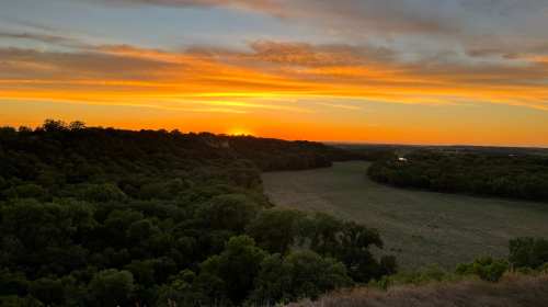 A vibrant sunset over a lush green landscape, with rolling hills and a river in the distance.