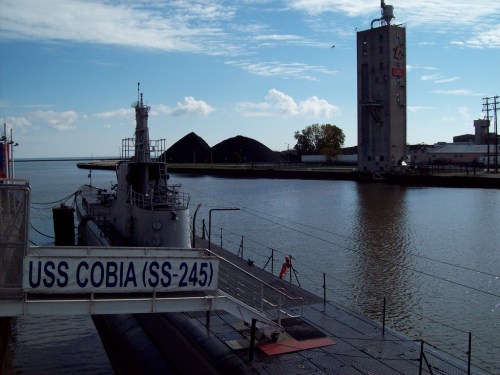 A submarine, USS Cobia (SS-245), docked near a coal tower and calm water under a blue sky.