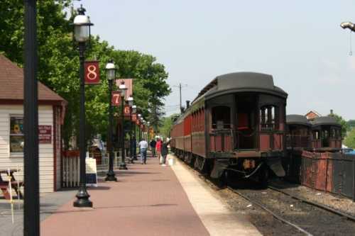 A vintage train at a station with passengers walking along the platform under clear skies and green trees.