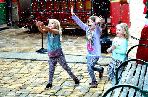 Three children joyfully play in falling snow, reaching out with their hands and smiling in a festive outdoor setting.