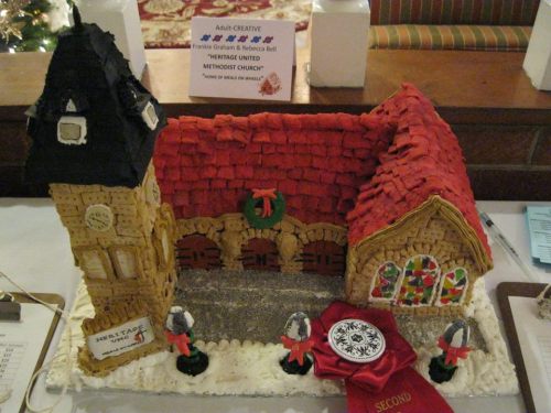 Gingerbread church with a red roof, clock tower, and festive decorations, displayed at a holiday event.