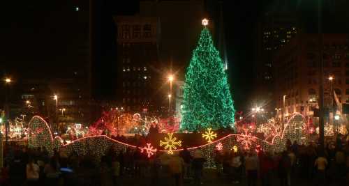 A brightly lit Christmas tree surrounded by festive lights and decorations, with a crowd enjoying the holiday atmosphere.