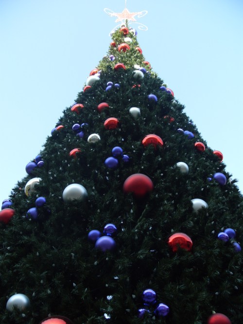 A tall Christmas tree adorned with red, silver, and purple ornaments against a clear blue sky.