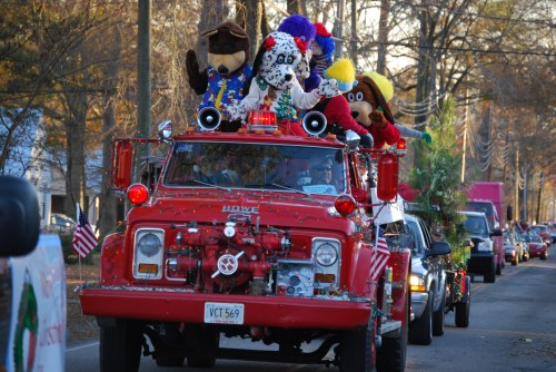 A festive red fire truck adorned with holiday decorations and plush characters, parading down a tree-lined street.