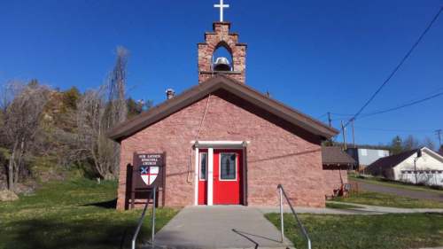 A small stone church with a red door and bell tower, surrounded by grass and trees under a clear blue sky.