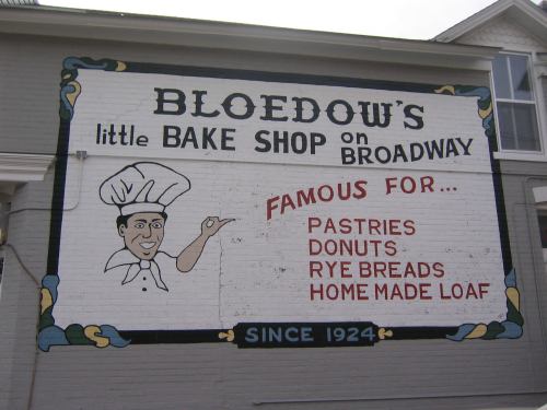 Mural of Bloedow's Bake Shop sign featuring a chef and listing pastries, donuts, rye breads, and home-made loaf since 1924.