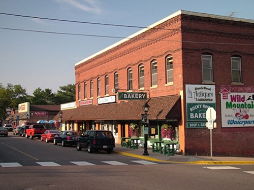 A quaint street scene featuring a brick building with shops, including a bakery, and parked cars along the road.