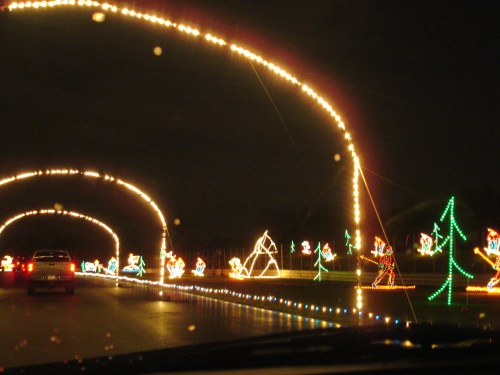 Colorful holiday lights arch over a road, featuring festive decorations like trees and animals against a dark sky.