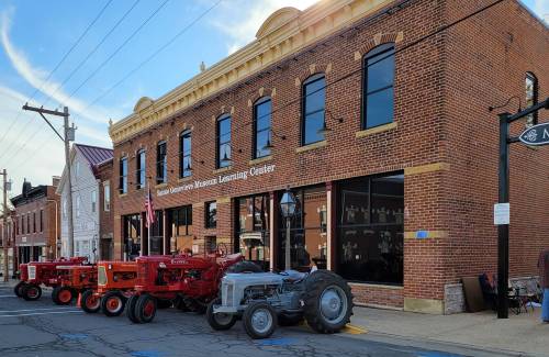 Historic brick building with large windows, featuring vintage tractors parked outside in a small town setting.