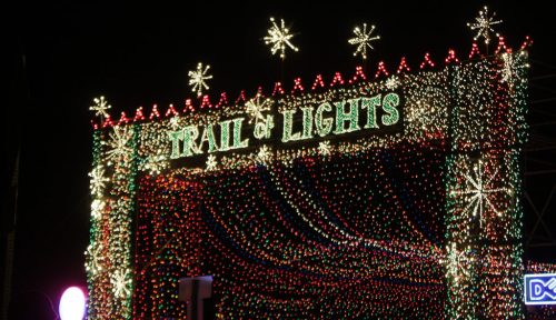 A festive display reading "Trail of Lights," adorned with colorful lights and snowflake decorations against a dark sky.