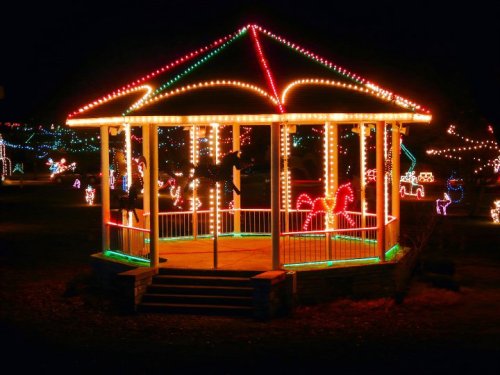 A brightly lit gazebo adorned with colorful holiday lights, surrounded by festive decorations in a dark night setting.
