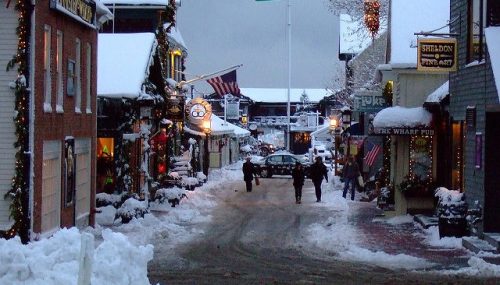 Snow-covered street lined with festive shops, American flag flying, and people walking in a winter wonderland.