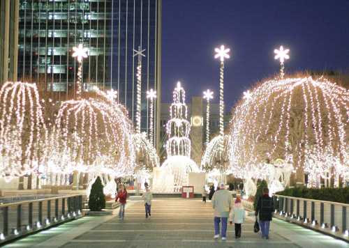 A festive walkway adorned with sparkling lights and star decorations, with people strolling in the evening.