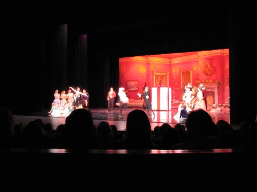 A theater stage with actors in period costumes performing in front of a colorful backdrop, with an audience in silhouette.
