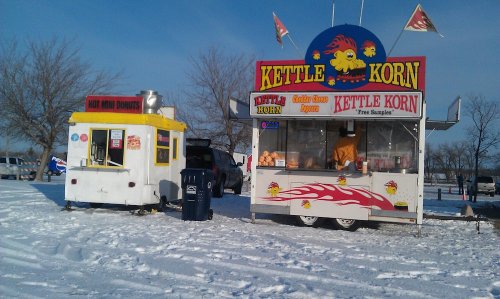 A snowy scene featuring two food trucks: one selling kettle corn and the other offering hot mini donuts.