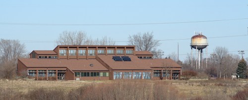 A modern brown building with solar panels, surrounded by trees, next to a water tower on a clear day.